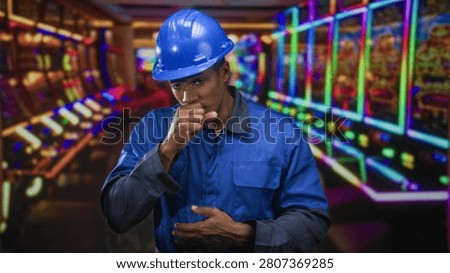 Man coughing into hand while clutching stomach, wearing blue hardhat and coveralls in casino slot machine aisle; workplace illness concern.