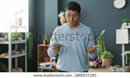 Man counts dollars while touching hair at a flower studio worktable with pots, vases, a roll of wrapping paper and potted plants visible; contemplation.