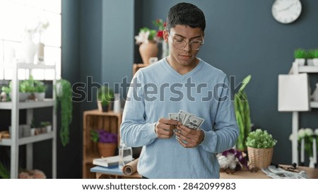 Man counts dollars while touching hair at a flower studio worktable with pots, vases, a roll of wrapping paper and potted plants visible; contemplation.