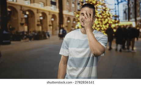 Man covering face with hand while smiling in a decorated building with blurred christmas tree and shoppers; embarrassed amusement.