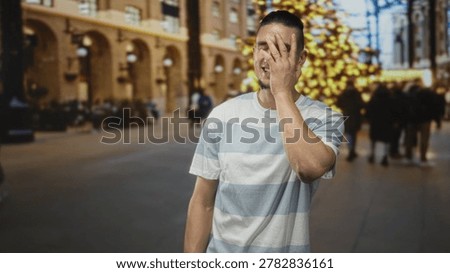 Man covering face with hand while smiling in a decorated building with blurred christmas tree and shoppers; embarrassed amusement.