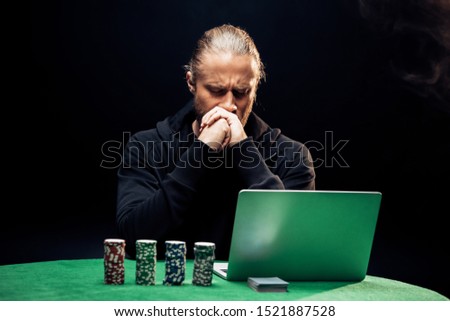 man covering face while using laptop near poker chips and playing cards on black with smoke