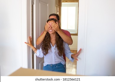 Man Covering His Girlfriend Eyes With Palms And Leading Her Into Their New Apartment. Family Couple Walking Through Doorway. Medium Shot. New Home Or Surprise Concept