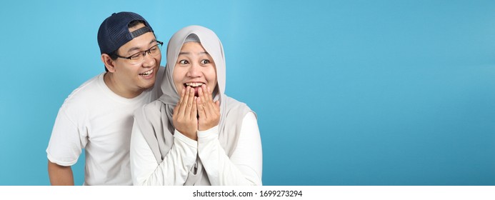 Man covering his mouth and whispering secret to his wife ear, Asian muslim woman looking shocked and excited, opening eyes and mouth widely with surprise, family concept
