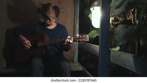 Man Cowboy On Dark Night With Acoustic Guitar Is Sitting On Porch Of Farmhouse, Playing An Instrument And Singing Songs. Concept Of Creativity, Hobbies, Lifestyle And Relaxation. Old, Retro Decor