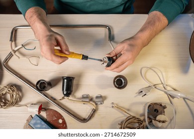 Man crafter disassembling wire system holder details of old vintage lamp, unscrewing internal parts of light bulb socket, removing screws with screwdriver tool, creative upcycling workshop process.