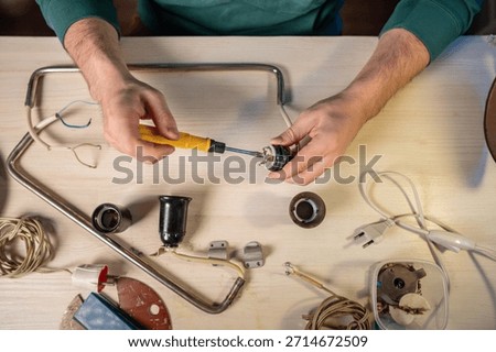Man crafter disassembling wire system holder details of old vintage lamp, unscrewing internal parts of light bulb socket, removing screws with screwdriver tool, creative upcycling workshop process.