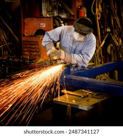 A man creating bright sparks while grinding metal in a machine shop. The man is wearing a long sleeve blue shirt and blue jeans