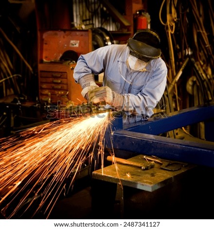 A man creating bright sparks while grinding metal in a machine shop. The man is wearing a long sleeve blue shirt and blue jeans