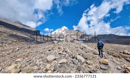 A man crossing a stony slope towards Mount Kazbeg in Caucasus, Georgia. Barren slopes below the snow-capped peak and the Gergeti Glacier. Tranquillity. Natural remedy. Massive glacier foot. Few cloud