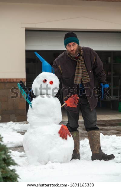man crouched next to a snowman figure of three\
large snow balls in the background of the garage. The snowman has a\
watering can and a\
brush\
\
