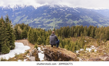 Man Crouching On A Rock. Foto From Behind. Man Is Looking To The Mountains In The Distance.