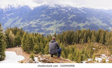 Man Crouching On A Rock. Foto From Behind. Man Is Looking To The Mountains In The Distance.