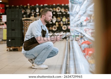 Man crouching while looking at products in refrigerated section of supermarket, holding tablet for convenience and information