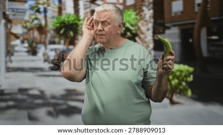 Man cupping ear while holding banana with right hand on a sunny street lined with palm trees and shops; absurdity confusion.