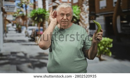 Man cupping ear while holding banana with right hand on a sunny street lined with palm trees and shops; absurdity confusion.