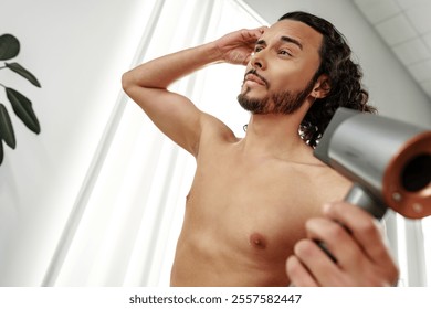 A man with curly hair styles himself in a bright room while using a hairdryer to enhance his look - Powered by Shutterstock