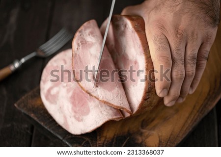 Man cuts ham sausage on cutting board. Knife is in hands of man, fork is on table. Thin slices of ham. Dark wooden table. Dark blurred background. Top view. Close-up. Soft focus. 