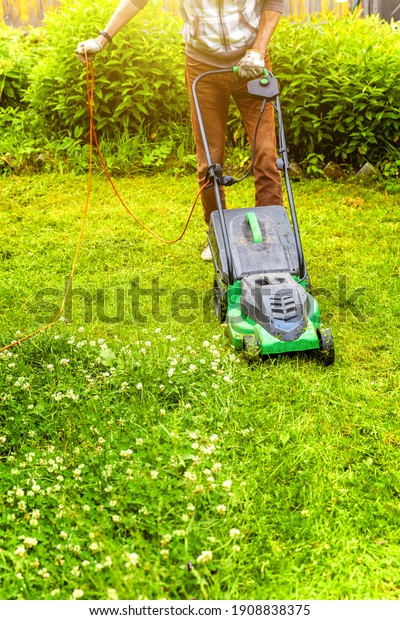 Man\
cutting green grass with lawn mower in backyard. Gardening country\
lifestyle background. Beautiful view on fresh green grass lawn in\
sunlight, garden landscape in spring or summer\
season