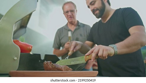 Man cutting meat at barbecue. Chef preparing food for family and friends. Father holding bottle of beer observing adult son prepare meal at home backyard grill - Powered by Shutterstock