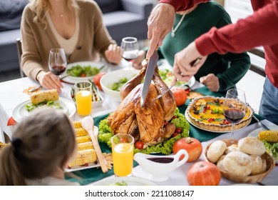 Man Cutting Turkey Near Blurred Family And Tasty Thanksgiving Dinner