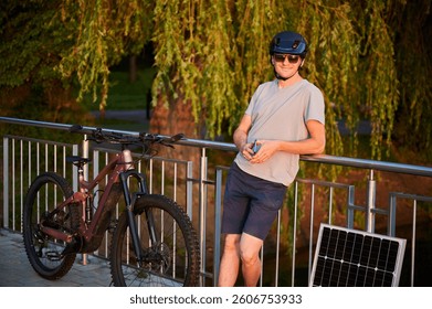 Man cyclist using solar panel for charging electric mountain bike outdoors. Happy guy in helmet, sunglasses leans against railing, holding phone, enjoying peaceful, sunlit setting near lush greenery.