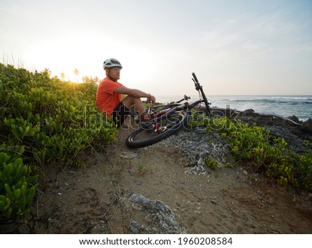 Man cyclist wearing white helmet and orange t-shirt seating with a mountain bike, at ocean shoreline. Rocky beach.