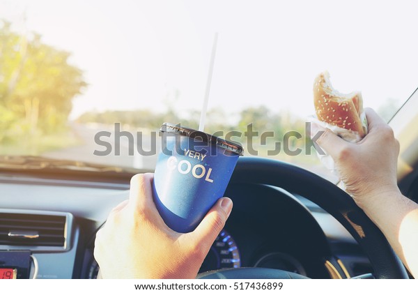 Man is dangerously eating hot dog and cold drink\
while driving a car