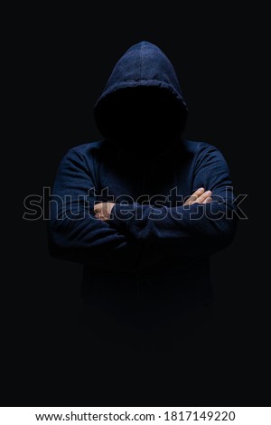 Man in dark blue hoodie, face hidden in shadow, unrecognizable, crossed arms, alone in dark 