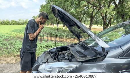Man in dark clothes examining engine of broken-down car on rural roadside with a worried expression, surrounded by fields and trees, symbolizing unexpected travel issues, car trouble concept