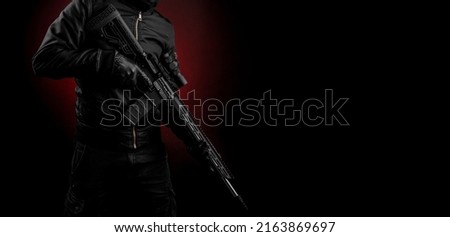 A man in dark clothes holds an automatic carbine in his hands. The face is covered with a mask, hands are in gloves. A figure with a weapon on a dark background with a red light spot.