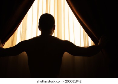 Man In Dark Room Opens Curtains On Window To The Morning Sunlight