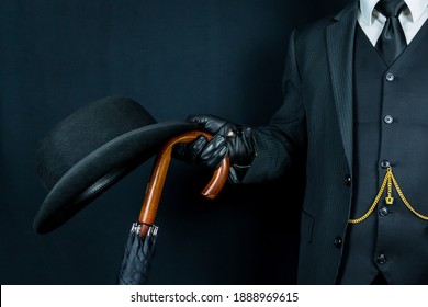 Man in Dark Suit and Leather Gloves Holding Bowler Hat and Umbrella on Black Background. Concept of Classic and Eccentric British Gentleman Stereotype. Retro Style and Vintage Fashion.