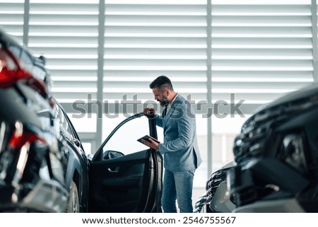 A man in a dealership inspects a car while holding a digital tablet, reflecting a thorough and detailed approach to evaluating the vehicle's features and condition before making a purchase.