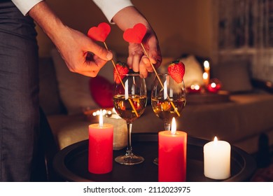 Man Decorating Table Setting For Valentines Day Celebration At Home Putting Hearts In Glasses With Champagne And Strawberries On Top By Candles On Table. Night Date