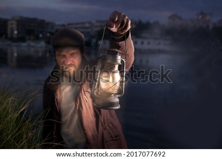a man in defocus with an evil face with a beard and in vintage clothes holds a kerosene lamp in his hands. It's dark around, sea background