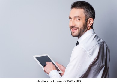 Man With Digital Tablet. Rear View Of Happy Mature Man In Shirt And Tie Working On Digital Tablet And Looking Over Shoulder While Standing Against Grey Background