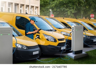 A man with a digital tablet stands next to yellow electric delivery vans at electric vehicle charging stations