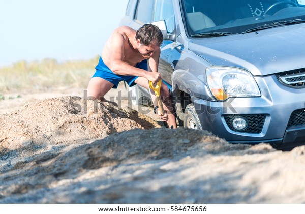 man digs a car stuck in the sand, throwing sand\
shovel out from under the\
car