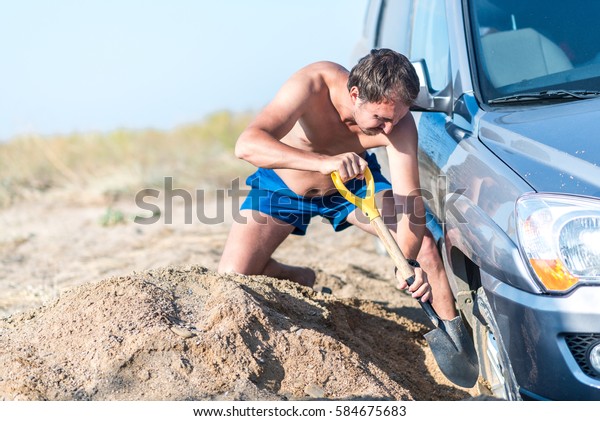 man digs a car stuck in the sand, throwing sand\
shovel out from under the\
car