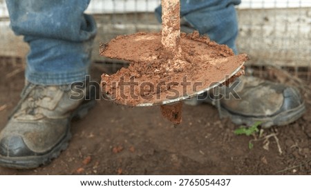 A man digs a hole in the ground for a post using a hand auger. A close-up of the man's feet in boots and the cutting edge of the auger with the soil removed.                               