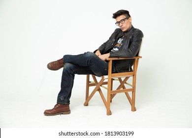 Man In A Director's Chair On A White Background In The Studio, A Man In A Black Leather Jacket, Jeans And Glasses. Modern Film Art