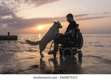 Man with disability, electric wheelchair user petting his dog and enjoying a sunset on the beach. People and animal friendship concept. - Powered by Shutterstock