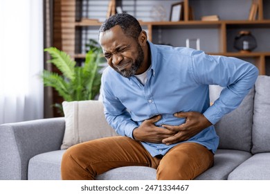 Man in discomfort, holding his stomach while sitting on a sofa, indicating possible pain or indigestion in a home setting. - Powered by Shutterstock