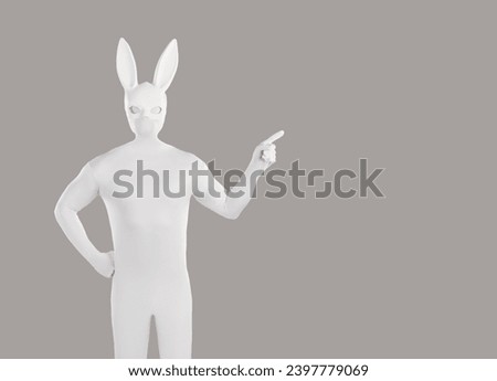 Man disguised in rabbit costume advertises something on blank grey copyspace background. Anonymous guy wearing white bodysuit and long eared playboy mask standing with hand on hip and pointing to side