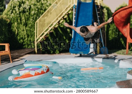 A man dives headfirst into a backyard swimming pool from a blue water slide. The sunny summer scene shows pool floats and play objects near the water.