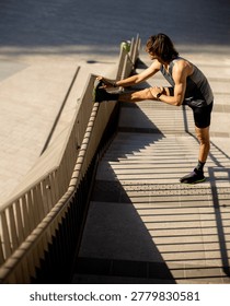 A man does a leg stretch while holding onto a railing. He is near stairs in an urban setting. Sunshine casts shadows on the ground, and it looks like a warm day.