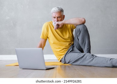 A Man Does Yoga And Fitness In The Gym With A Trainer Online From A Laptop. Senior Male Sports Coach On The Internet. Senior Yoga At Home Or In The Gym For Training. A Fifty-year-old Man With Gray