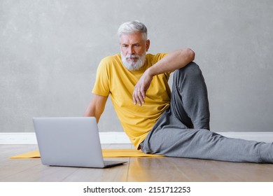 A Man Does Yoga And Fitness In The Gym With A Trainer Online From A Laptop. Senior Male Sports Coach On The Internet. Senior Yoga At Home Or In The Gym For Training. A Fifty-year-old Man With Gray