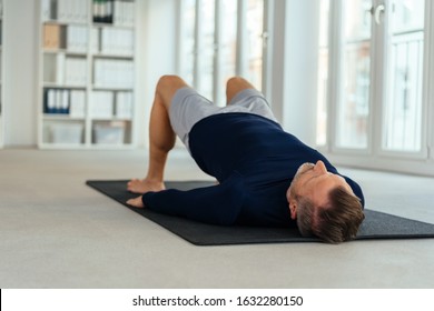 Man Doing Bridging Exercise, Lying On His Back On Black Mat In Empty Office Interior. Viewed From Floor Level From His Head